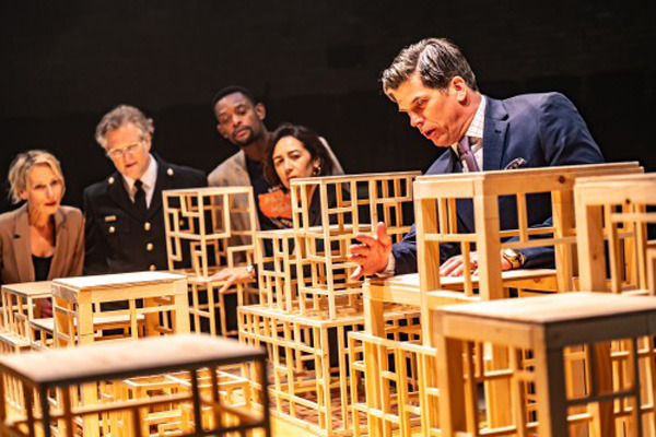 Five actors in costume surrounding a group of abstract wooden cube structures on a stage. They are engaged in an intense moment of performance or rehearsal, with dramatic lighting highlighting their expressions.